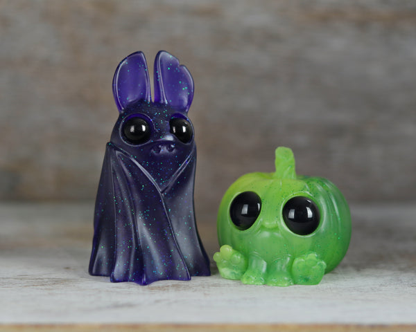 A small, glittery purple bat resin figure with big black eyes stands beside the adorable green Kabocha Pupkin—an endearing pumpkin-shaped creature with wide eyes and tiny feet—on a wooden surface, perfectly capturing the enchanting atmosphere of the Nightshade Belfry & Kabocha Pupkin PRE-ORDER collection from Bindlewood Shop amid a softly blurred background.