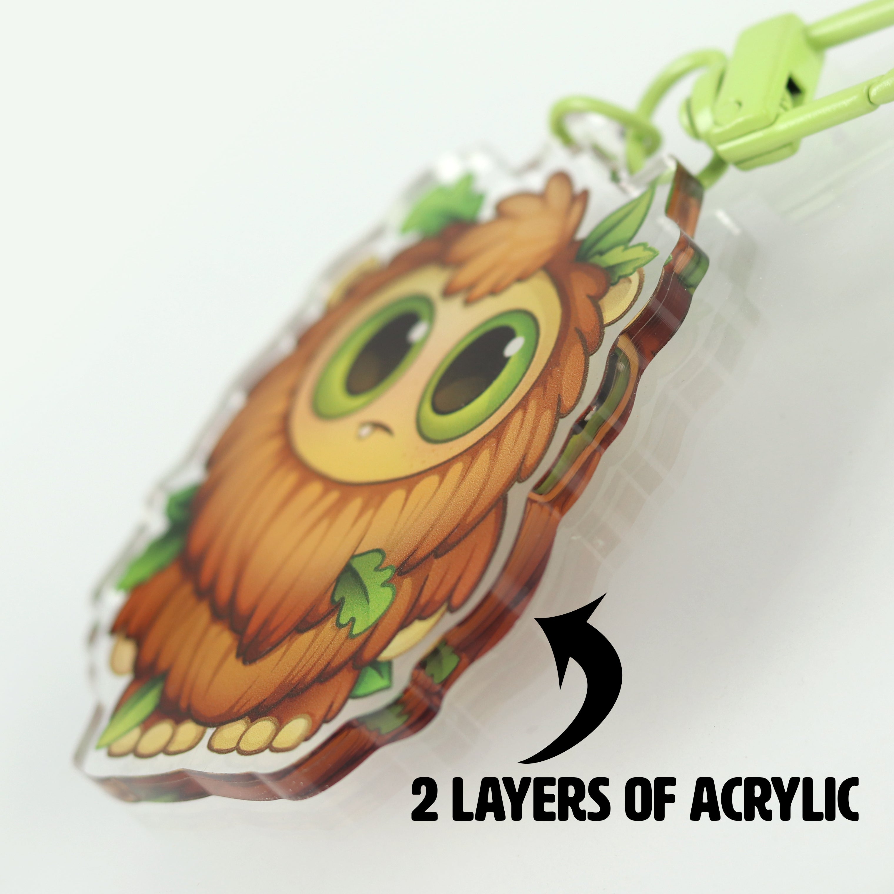 A close-up of the Toastboy Keychain by Bindlewood Shop, a cute cartoon charm with leafy accents and big green eyes, inspired by Chris Ryniak. Made from two layers of acrylic (as noted with an arrow on the side).