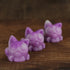 Three small, purple Studgy soaps shaped like cats with large eyes, hand cast with care and attention to detail by Chris Ryniak, are arranged in a diagonal line on a wooden surface. The brown blurred background adds warmth, complementing the whimsical charm of these unique creations.