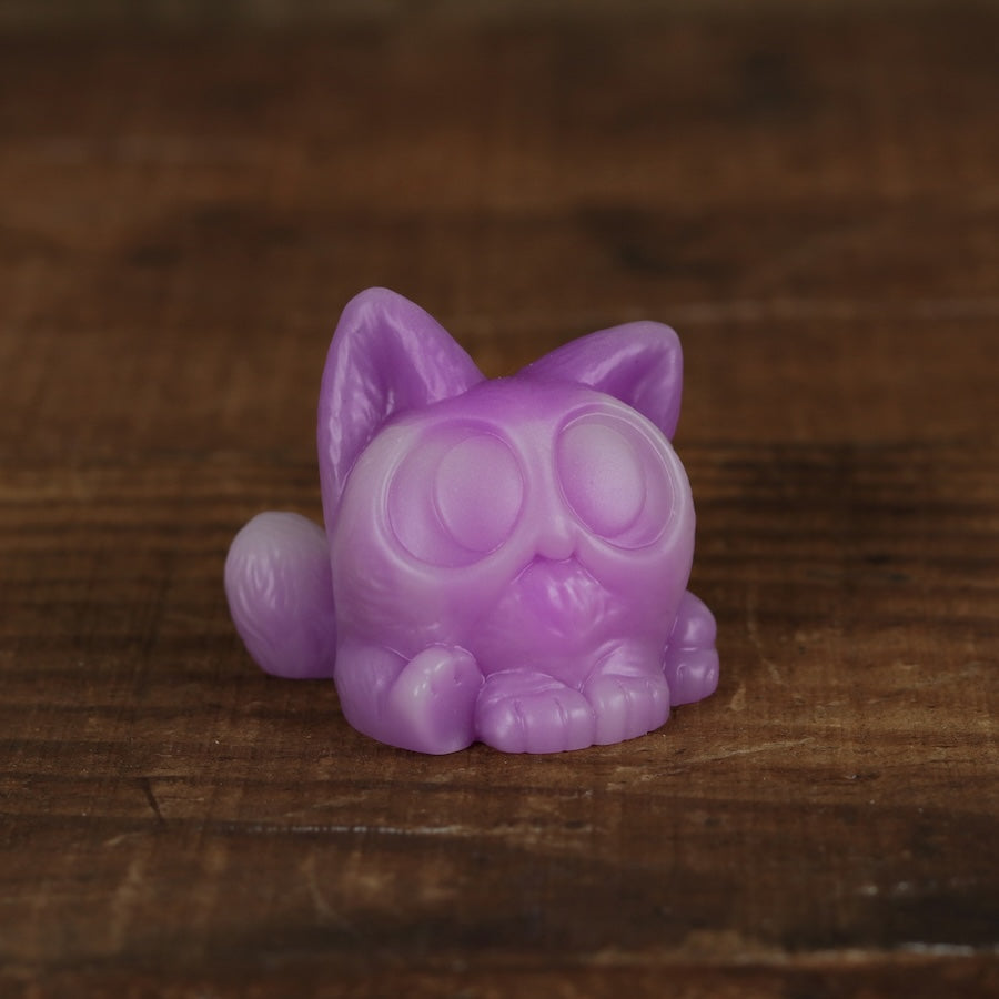 A small, purple Studgy figurine by Chris Ryniak, hand-cast from resin and featuring large round eyes in a cat shape, sits on a wooden surface.