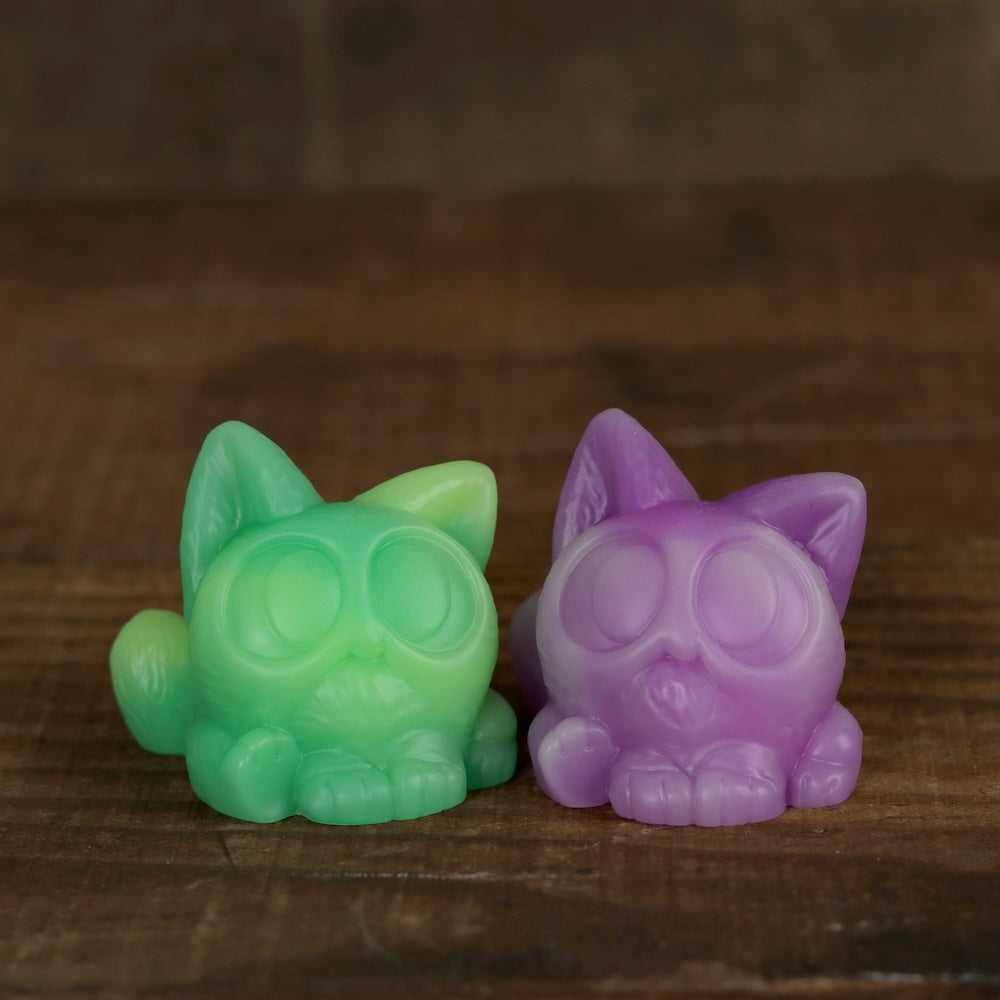 Two small, round Studgy cat figurines from Chris Ryniak sit side by side on a wooden surface. Hand cast and featuring exaggerated eyes, one figurine is green and yellow while the other is pink and purple. The background shows blurred brown wood.