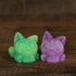 Two small, round Studgy cat figurines from Chris Ryniak sit side by side on a wooden surface. Hand cast and featuring exaggerated eyes, one figurine is green and yellow while the other is pink and purple. The background shows blurred brown wood.