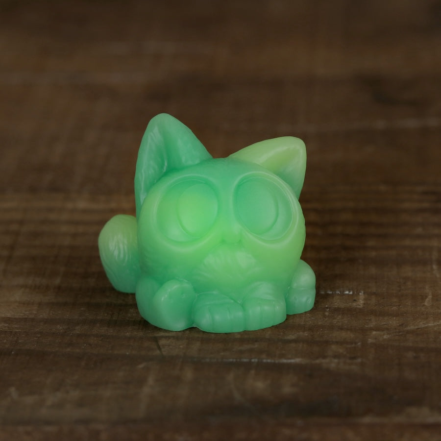 A small green Studgy cat figurine by Chris Ryniak, featuring big round eyes and hand-cast resin craftsmanship, sits on a wooden surface.