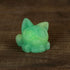 A small green Studgy cat figurine by Chris Ryniak, featuring big round eyes and hand-cast resin craftsmanship, sits on a wooden surface.