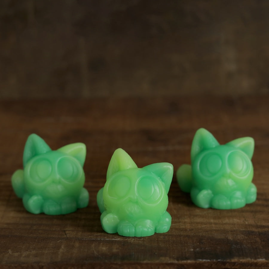 Three small, green, cat-shaped Studgy figurines with large round eyes sit on a wooden surface against a softly blurred brown background. These charming resin figures are hand cast and designed by artist Chris Ryniak.