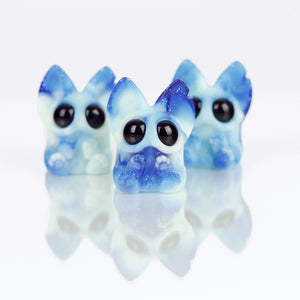 Three small, blue and white Sunshower Beans figurines by Chris Ryniak, with large black eyes and pointy ears, are displayed on a reflective surface. These adorable collectibles feature a cute, cartoon-style design.