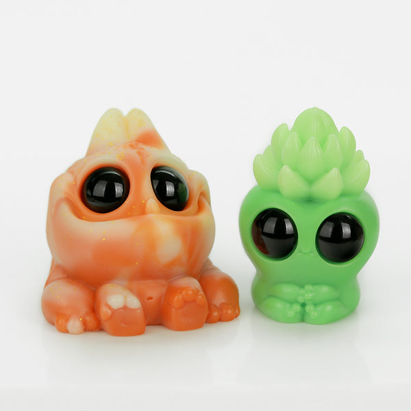 The Bindlewood Shop’s Thai Tea Tank & Matcha Latte Archie Set features two playful resin figures—an orange, frog-like Thai Tea Tank and a cactus-headed green Matcha Latte Archie—both with big black eyes and bright colors on a white surface.