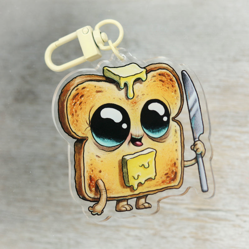 The Toastboy Keychain by Bindlewood Shop is a cute acrylic charm of a smiling toast slice with shiny eyes, melting butter, a little knife, and playful cartoon arms and legs—perfect for adding whimsy to your keys.