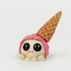 A cute, small figurine by Chris Ryniak with a round, white body, big black eyes, and tiny paws. The Single Scoop Turtball has pink fabric draped over its head and a waffle ice cream cone placed upside down on top of it, resembling a whimsical hat. This delightful piece debuted at the Five Points Festival.