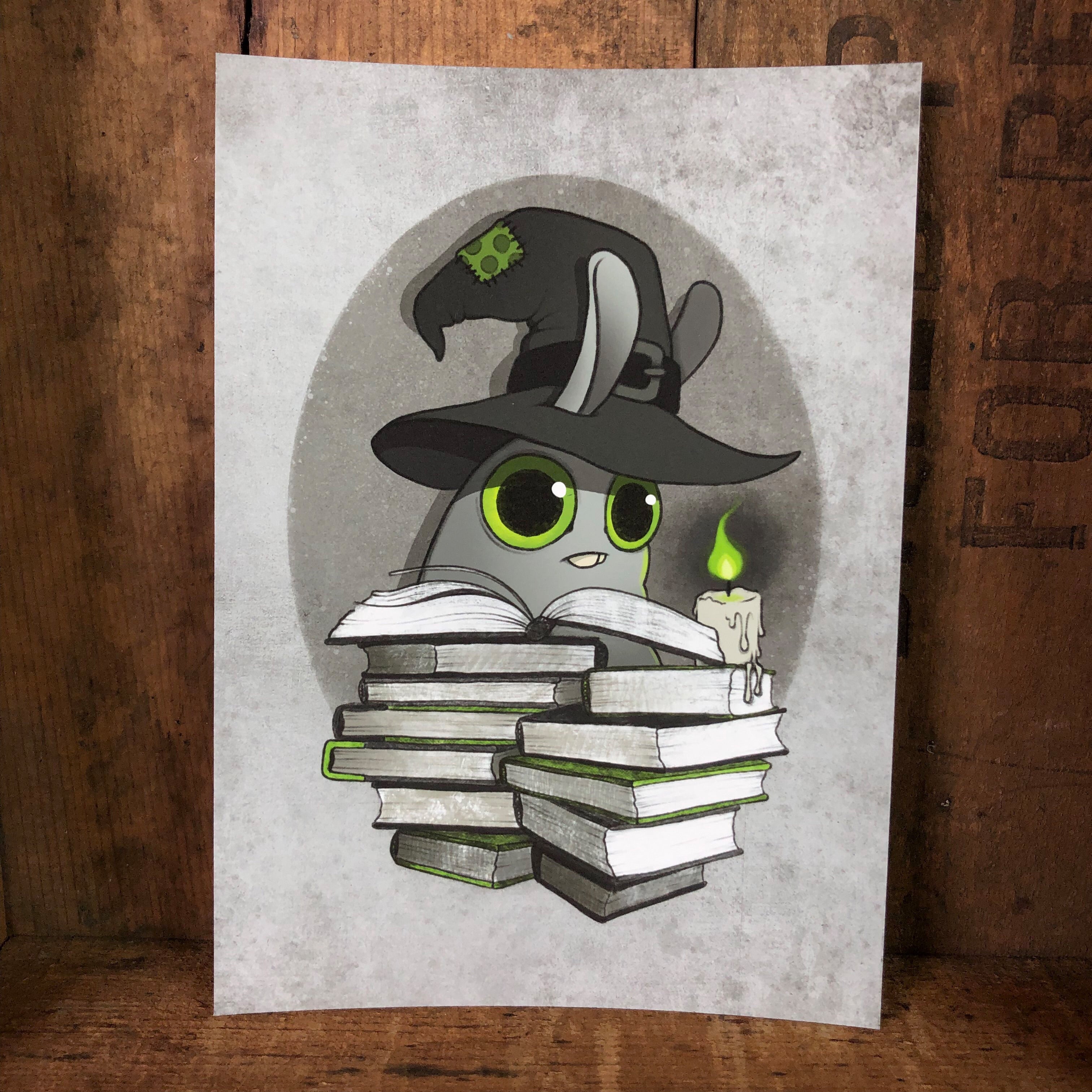 The "Witchbunny" Print by Amanda L Spayd features a cartoon rabbit with big green eyes and a black witch hat holding a glowing green candle behind books, set against a textured gray background on archival fine art paper.