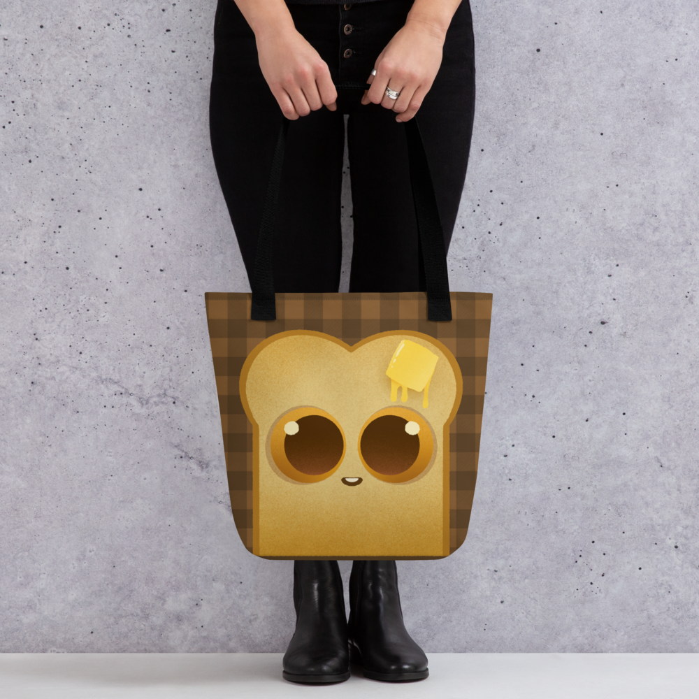 A person holds the Toast-Bag Tote by Bindlewood Shop, a durable bag with a cute cartoon bread slice (big eyes, melting butter) on a brown checkered background. The person wears black pants and boots.