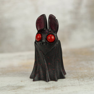 Holiday Belfry (Red) by Bindlewood Shop is a small, stylized bat figurine with large red eyes, a black body, and berry-red speckles. Designed by Amanda Louise Spayd, it stands upright with wings wrapped around its body.