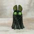 A small, black Holiday Belfry (Green) resin figure by Bindlewood Shop stands on a light wooden surface; it features green speckles, large round green eyes, and tall ears against a blurred neutral background.