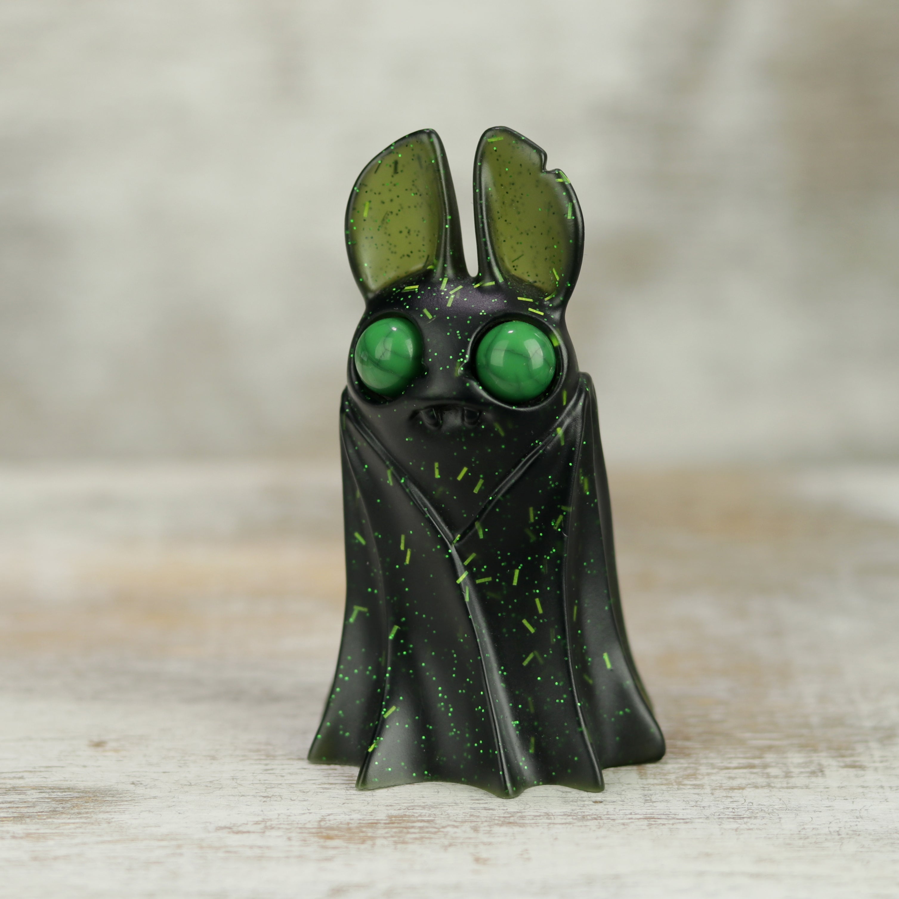 A small black resin bat figure with large green eyes and ears, accented by green speckles, stands on a wooden surface—a charming Holiday Belfry (Green) by Bindlewood Shop.