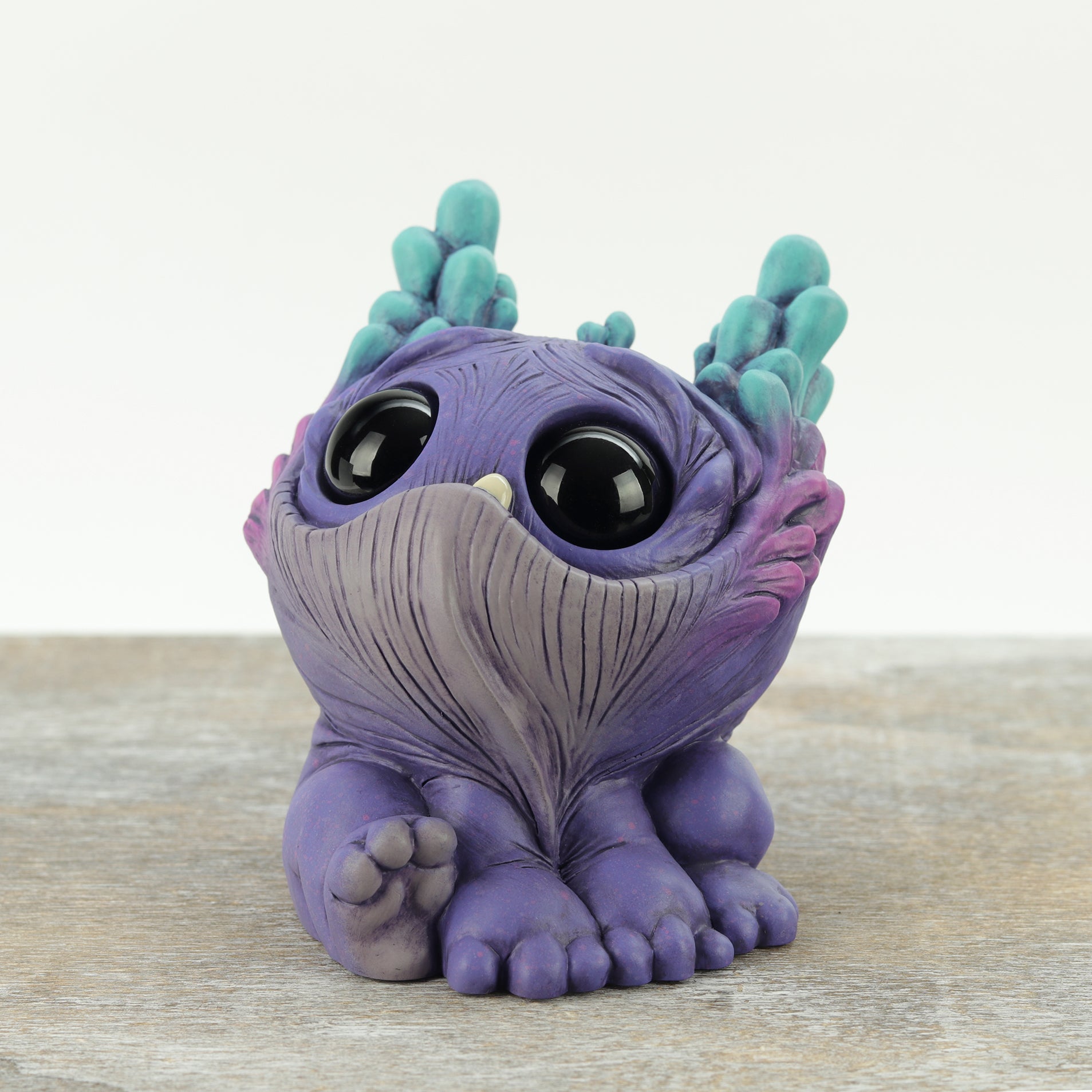 Dewlap by Chris Ryniak: a small, whimsical purple creature figurine with large black eyes, teal horns, wrinkled texture, and tiny feet sits on a wooden surface against a light background.