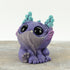 Dewlap by Chris Ryniak: a small, whimsical purple creature figurine with large black eyes, teal horns, wrinkled texture, and tiny feet sits on a wooden surface against a light background.