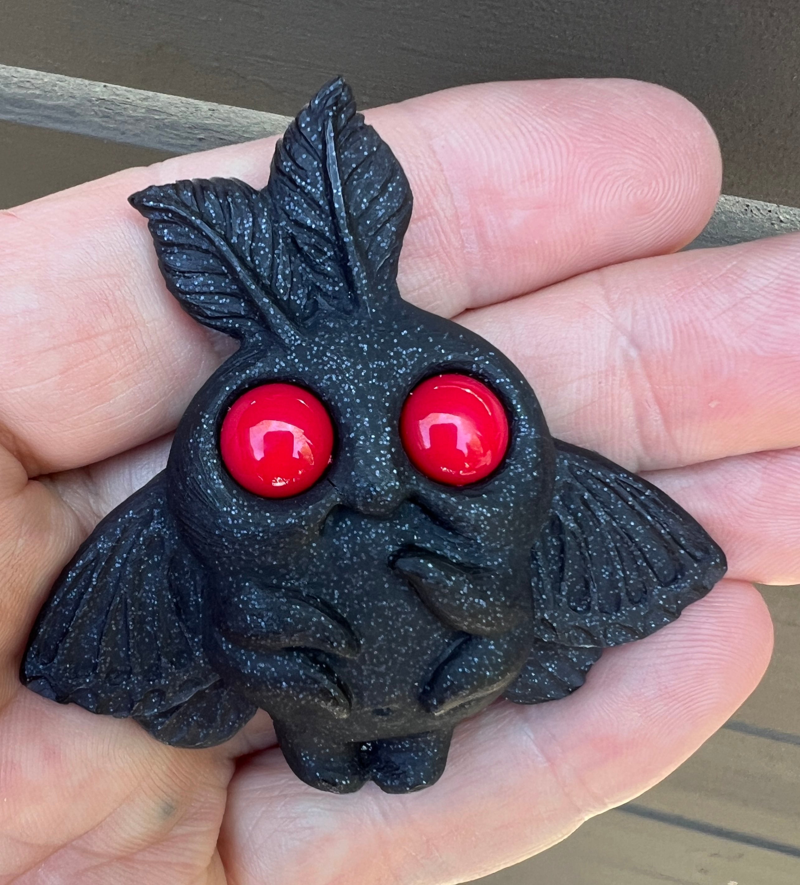 A person holds the "Mothman Magnet" by Chris Ryniak—a small black resin moth-like figurine with large red eyes, leaf-shaped ears, and scalloped wings—against a brown background.