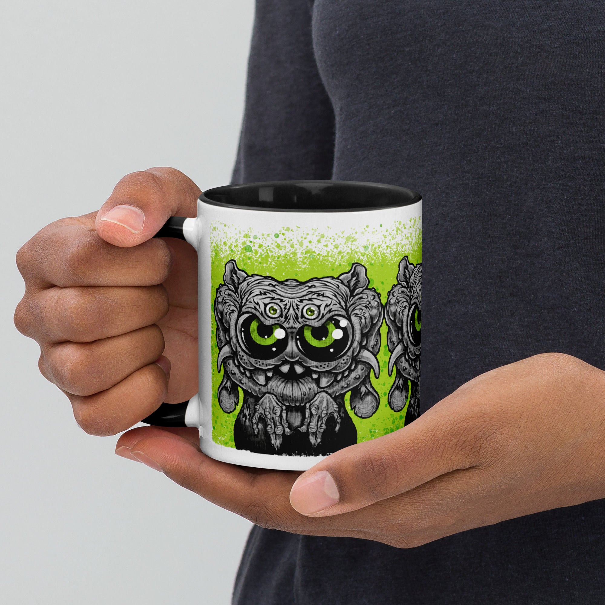 A person holds the Eyegurr Mug by Bindlewood Shop—a ceramic mug featuring a black-and-white cartoon monster with large, vivid green eyes and intricately detailed fur, set against a green background. This whimsical design is perfect for fans of collectible mugs and reminiscent of Chris Ryniak’s signature monster artwork.