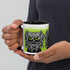 A person holds the Eyegurr Mug by Bindlewood Shop—a ceramic mug featuring a black-and-white cartoon monster with large, vivid green eyes and intricately detailed fur, set against a green background. This whimsical design is perfect for fans of collectible mugs and reminiscent of Chris Ryniak’s signature monster artwork.