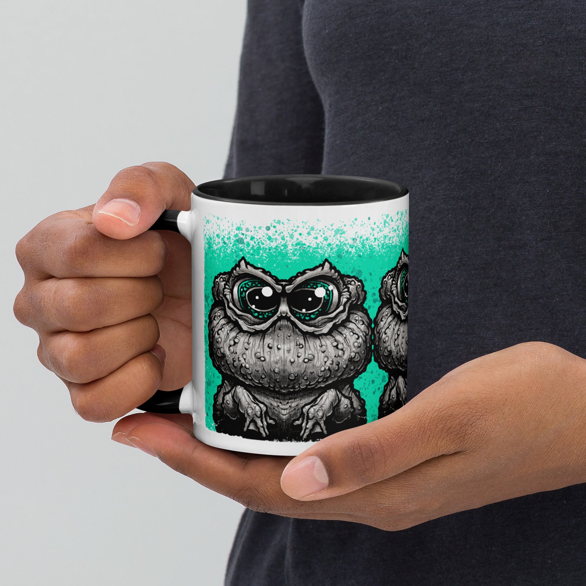 A person holding the Tode Mug by Bindlewood Shop—a white ceramic mug with a black interior, showcasing a detailed illustration of a cartoon owl with large eyes on a teal background. This collectible art mug adds charm and personality to any drinkware collection.
