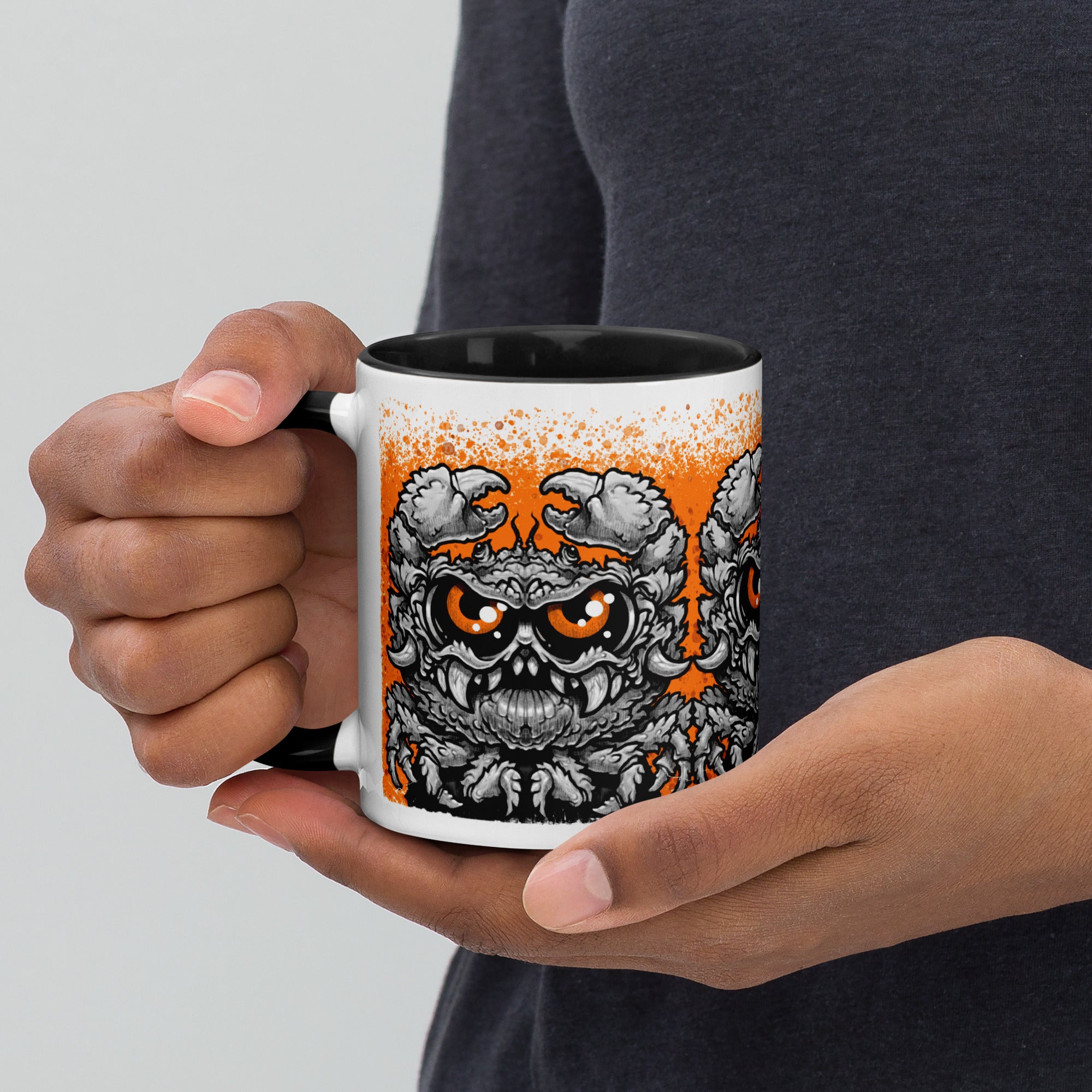 A person holds the Krabb Mug by Bindlewood Shop, a ceramic mug with a black handle featuring a detailed and menacing monster face illustration in black, gray, and orange tones against a white background—reminiscent of the signature style found in Chris Ryniak’s monster mugs.