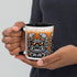 A person holds the Krabb Mug by Bindlewood Shop, a ceramic mug with a black handle featuring a detailed and menacing monster face illustration in black, gray, and orange tones against a white background—reminiscent of the signature style found in Chris Ryniak’s monster mugs.