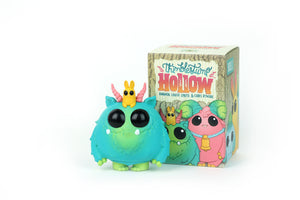 A cute blue and green monster figurine with big black eyes, pink horns, and a small yellow creature on its head stands next to a Bindlewood Shop Thimblestump Hollow Series 2 - Single Blind Box, featuring collectible character art.