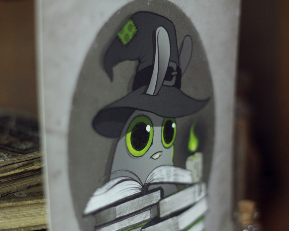 The "Witchbunny" print by Amanda L Spayd showcases a gray character with large green eyes, adorned in a witch hat, reading a book while holding a glowing candle. This fine art print is set against a soft, blurred background of visible books and captures the whimsical essence characteristic of Amanda L Spayd's style on archival paper.