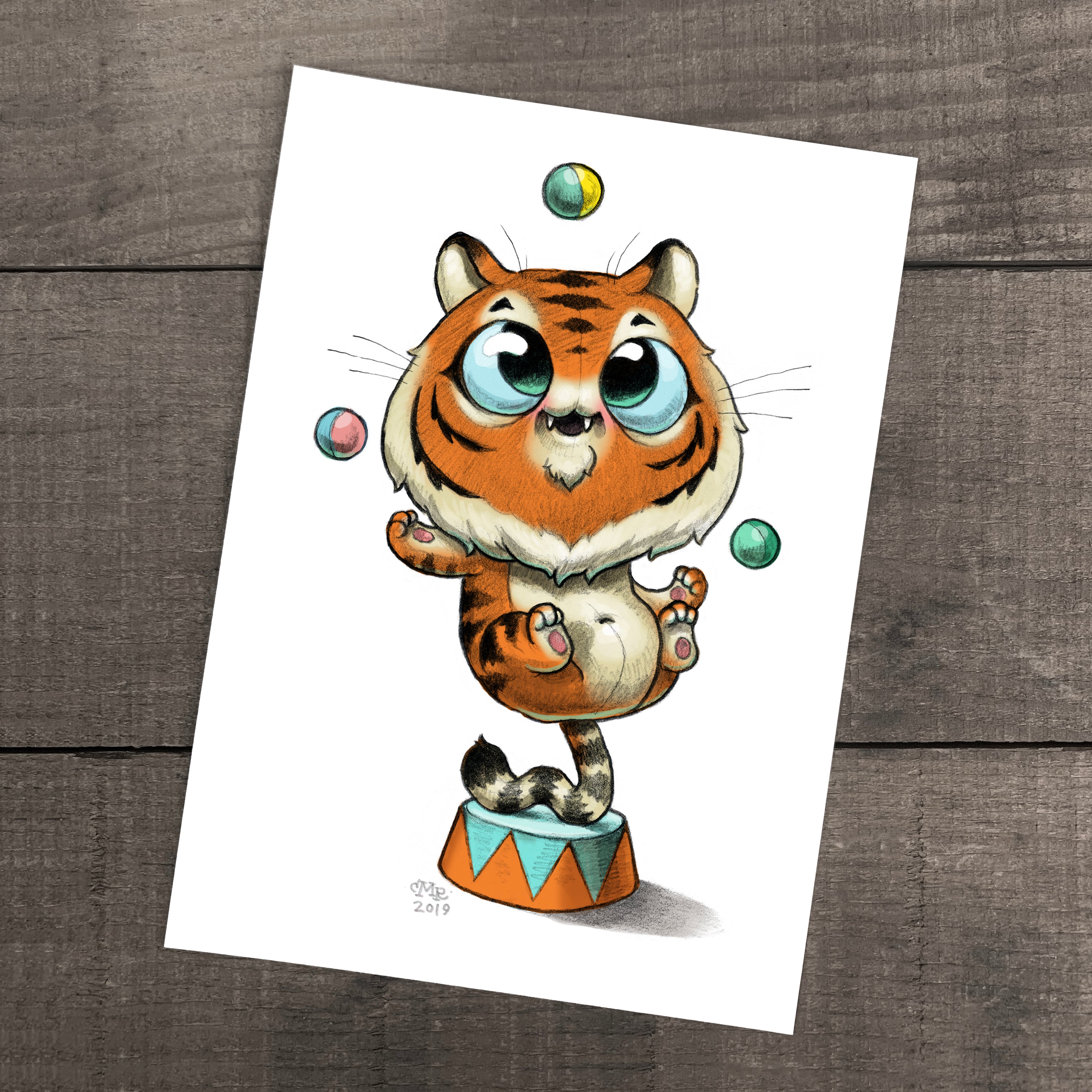 The Juggle Tiger Print by Chris Ryniak features a cute cartoon tiger with big blue eyes balancing on a circus stand and juggling four colorful balls, printed on archival paper and displayed atop a wooden surface.