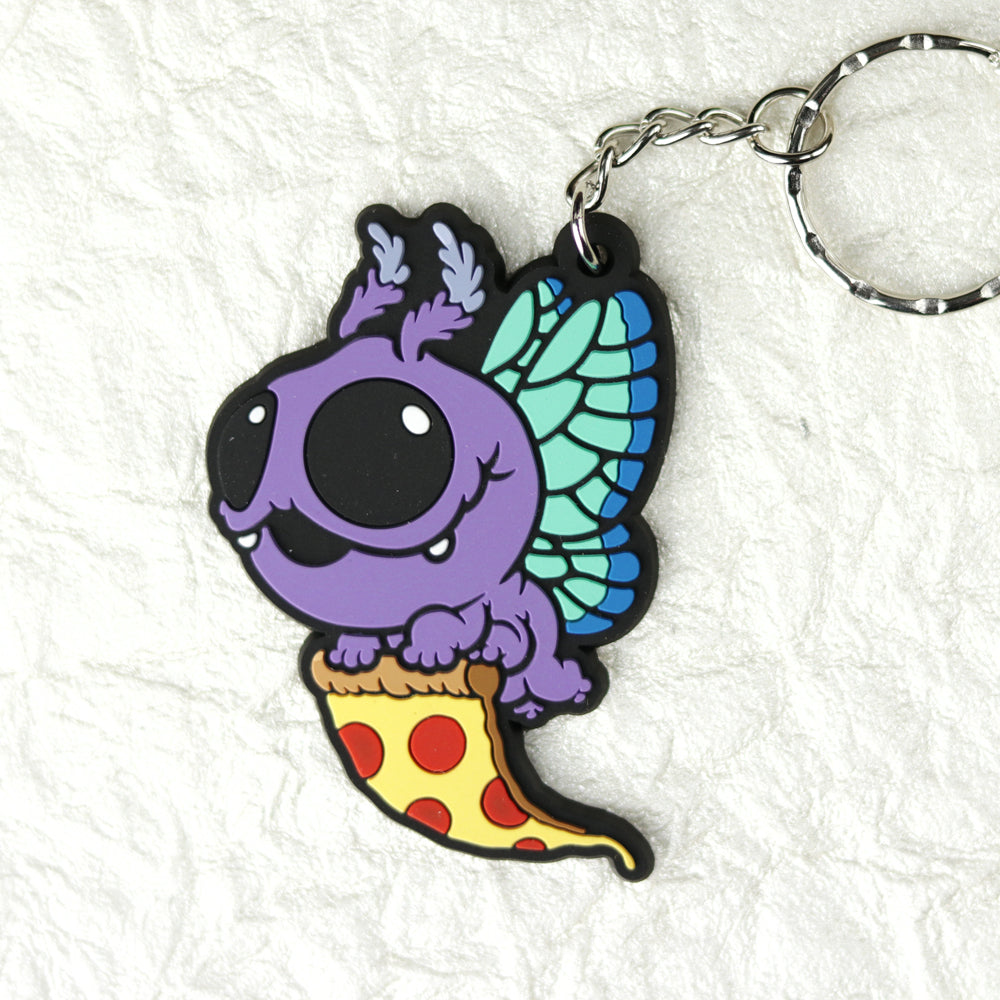 The Mothpup Keychain from Bindlewood Shop is a cute PVC rubber accessory featuring the purple cartoon bug from Thimblestump Hollow with big eyes and blue-green wings, riding a slice of pepperoni pizza on a textured white background.