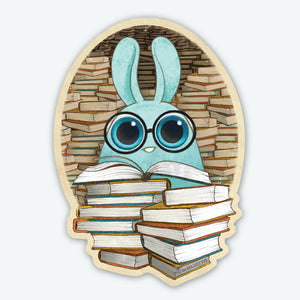 A cute blue Dust Bunny with big round glasses sits behind a stack of books, with more in the background on the Bindlewood Shop's 3-inch "So Many Books" vinyl sticker—perfect for Amanda Louise Spayd fans.