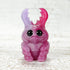 The MISFIT Sparkleberry Staggle by Bindlewood Shop is a small pink and purple resin figure with big black eyes and colorful pincers, shown on a white textured surface—a fun addition to any collection.