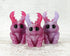 Three glossy purple and pink MISFIT Sparkleberry Staggle resin figures by Bindlewood Shop, featuring large black eyes and vibrant horns, stand on a white wooden surface against a light brick background.