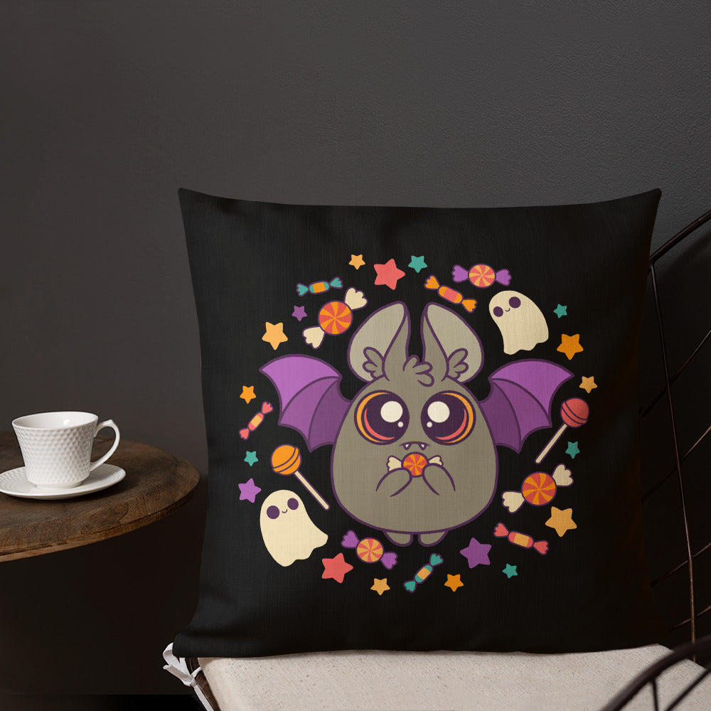 The Bindlewood Shop's Candy Bat Throw Pillow features Amanda Louise Spayd’s whimsical Candy Bat with purple wings, surrounded by colorful candies, stars, and two friendly ghosts. Ideal for Spooky Season decor, this premium-feel decorative pillow is beautifully displayed on a chair next to a small wooden table holding a white cup and saucer.