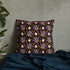 The Candy Bat Throw Pillow by Bindlewood Shop is a square cushion with a black background adorned with a vibrant pattern of cartoon owls and bats in shades of purple, grey, and yellow. Designed by Amanda Louise Spayd, this pillow is set against a concrete wall, resting on textured dark blue fabric with a plant visible on the left.
