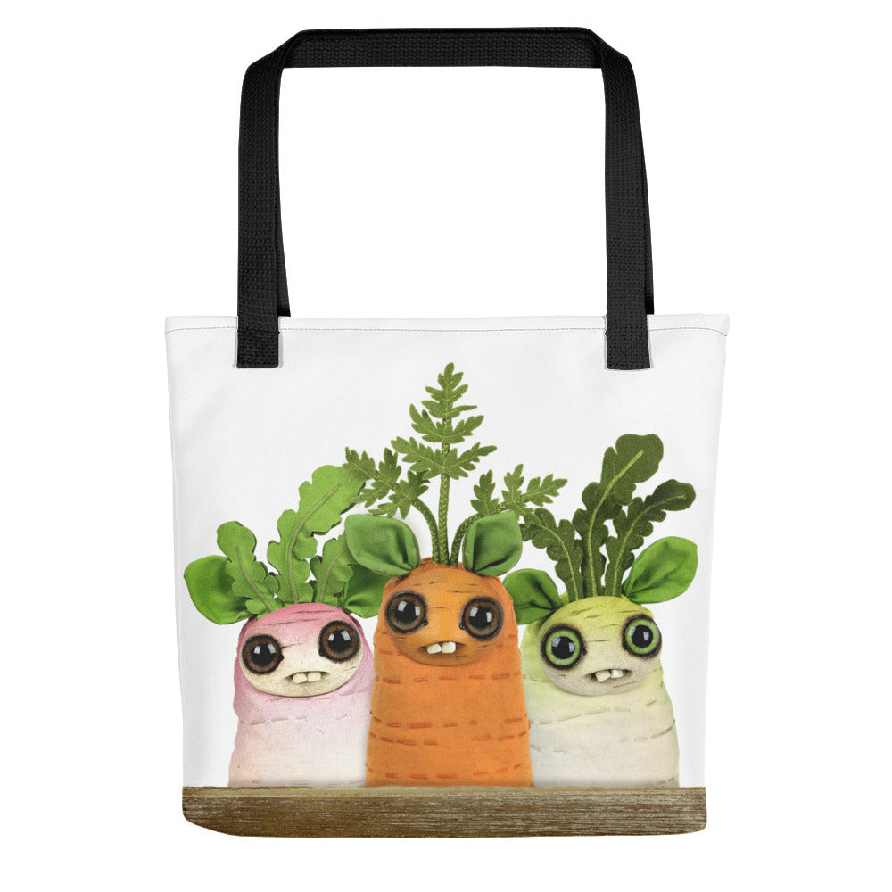 A white tote bag with black straps features a design of three cute, cartoon-style vegetables with big eyes and small smiles. From left to right, there are pink, orange, and green root vegetables with leafy green tops. Made from weather resistant fabric, this spacious and sturdy bag also showcases the Bindlewood Shop logo. Introducing the Farmer's Market Dust Bunnies Tote!