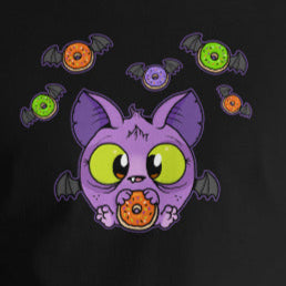 A person wearing a Bindlewood Shop Donut Bat Tee made from soft and lightweight combed and ring-spun cotton, featuring a colorful graphic of a cartoonish purple cat with big green eyes, surrounded by various candies. The individual is standing against a plain white background, with their hands relaxed by their sides.