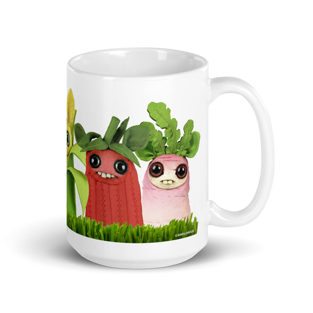 Introducing the Farm Market Dust Bunnies Mug by Bindlewood Shop—a white ceramic mug adorned with colorful, smiling plush vegetables featuring large eyes. From left to right, you'll find a yellow corn with a green husk, a red radish with green leaves, and a pink beet also with green leaves, all standing on a lush patch of green grass. This delightful design is both microwave safe and dishwasher safe.